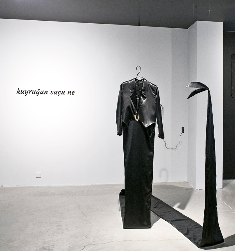 Black jacket with a long fabric tail on the floor in front of the wall text kuyruğun suçu ne