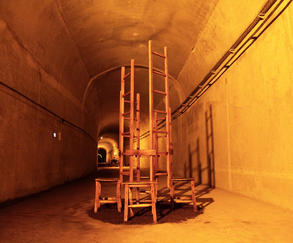 Sculpture Meeting: chairs stacked into a tall tower in an orange-lit tunnel