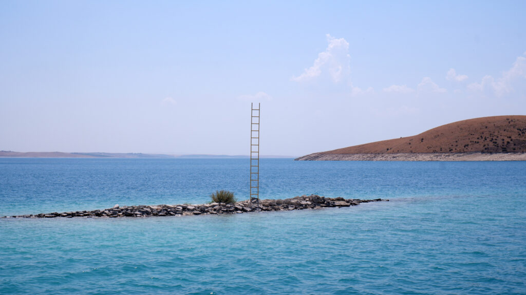 Tall ladder on a small stone islet in the dam lake, site-specific installation Rising