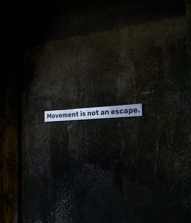 Text strip reading Movement is not an escape. on a dark wall, This is a Temporary Address